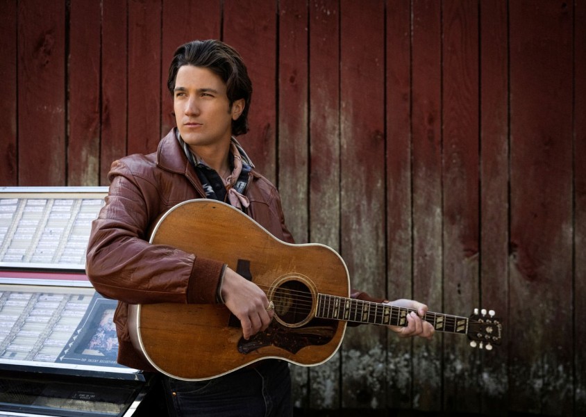 A man wearing a brown leather jacket holding an acoustic guitarx